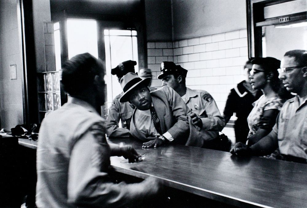 Black and white photograph of MLK sprawled across the police desk being restrained by two police officers as his wife and others look on. 