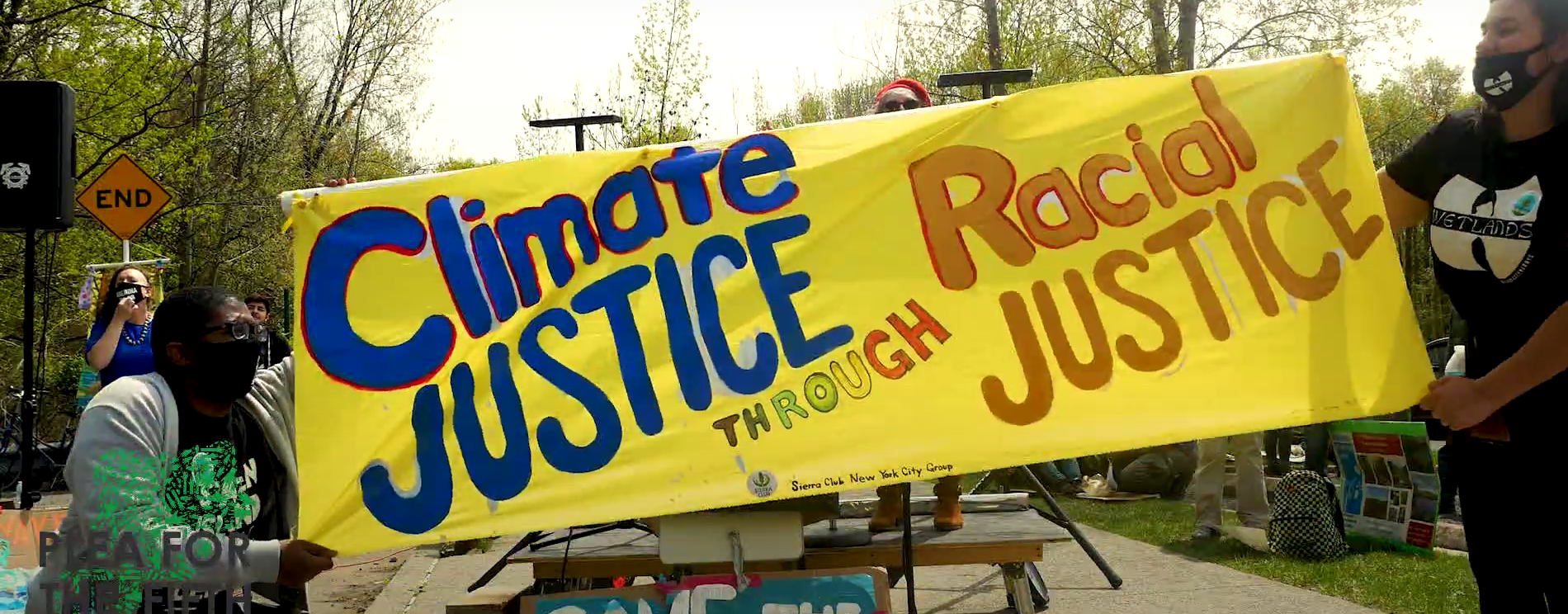 Two people holding either end of a yellow banner that reads Climate JUSTICE Through Racial JUSTICE in front of the stage.