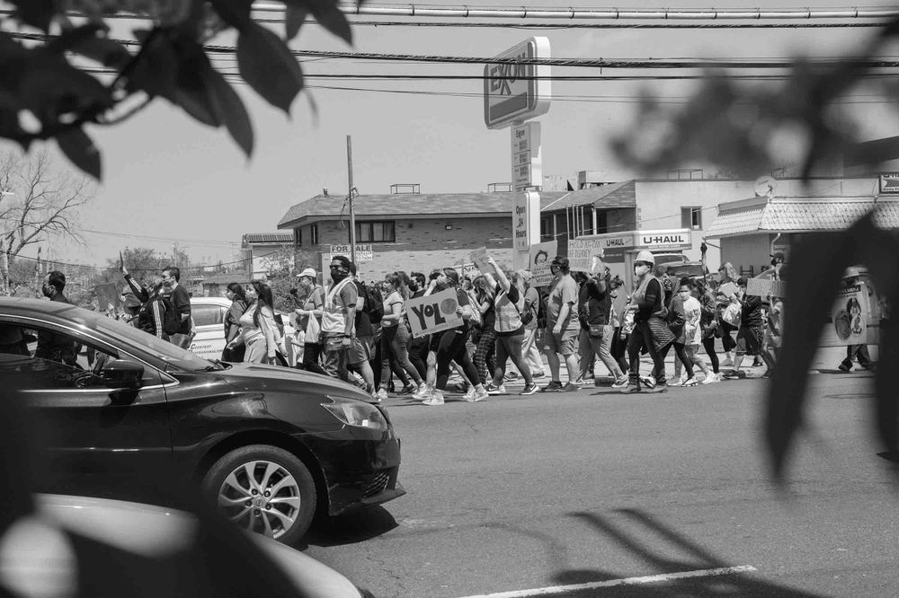 Black and white photograph of demonstrators marching on a street, Some are holsing signs, others have their hands raised in the air. 