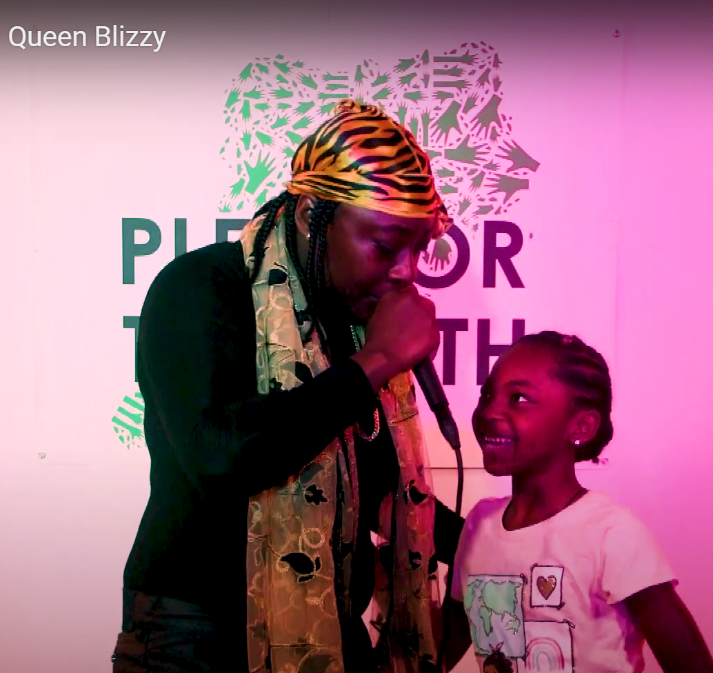 A women wearing all black wirh a leopard print headwrap and gold scarf singing to a girl in a pink t-shirt. PFT5's logo in partially visible in the background.