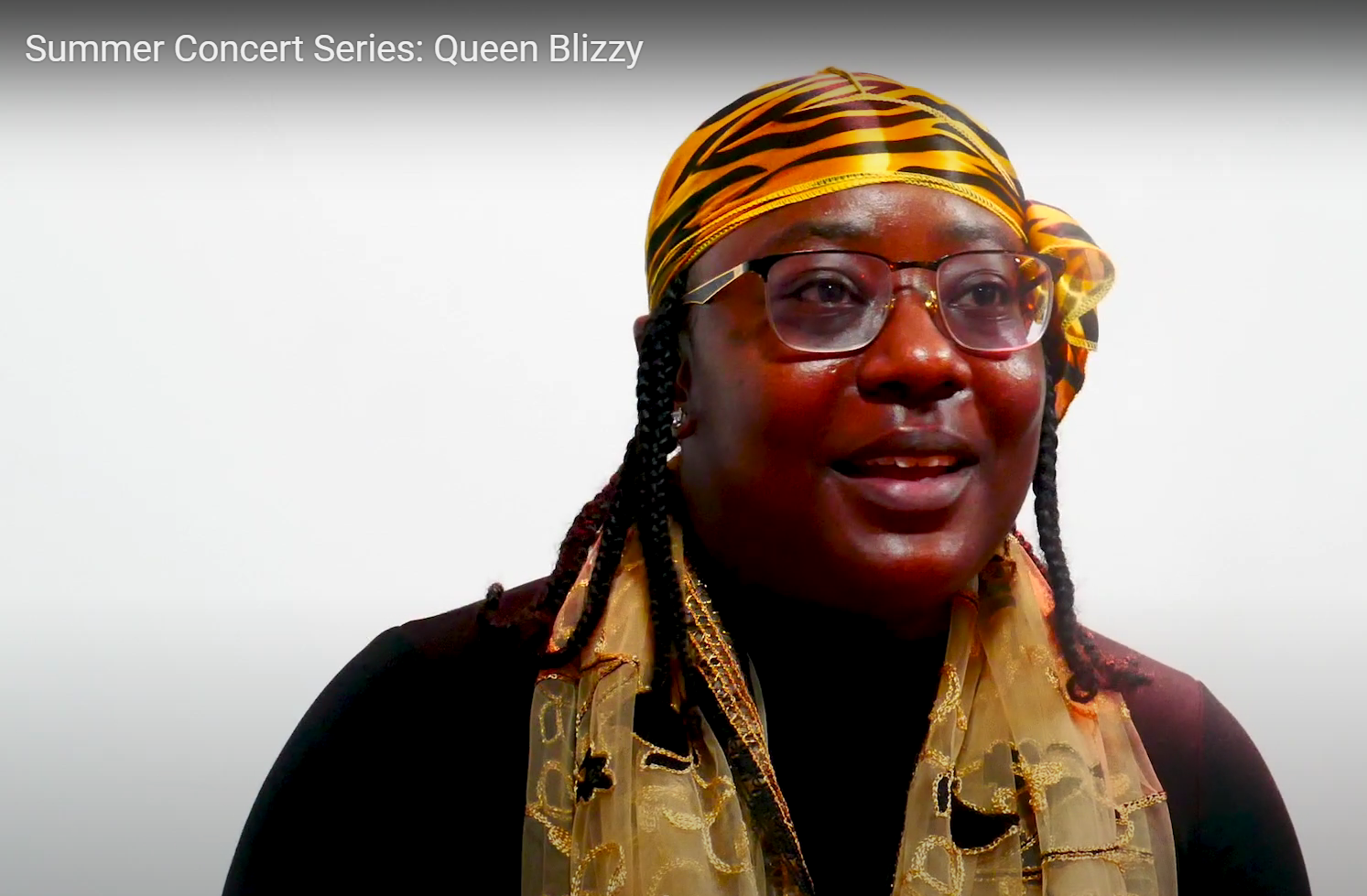 A black women wearing a black shirt with a leopard print headwrap and golden scarf.