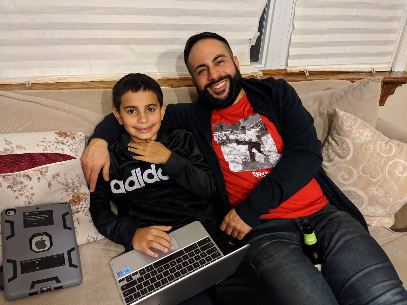 A man in a red t-shirt and cardigan embracing a child, his nephew on a couch. The child is wearing a black shirt and is holding a loptop.