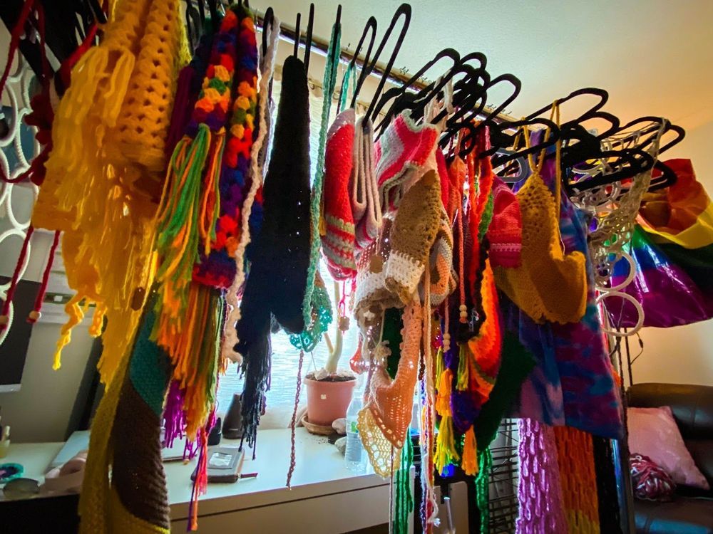 A clothes rack holding several crochet items in all different colors. Behind the clothes rack there is a white desk with a notebook and a plant.