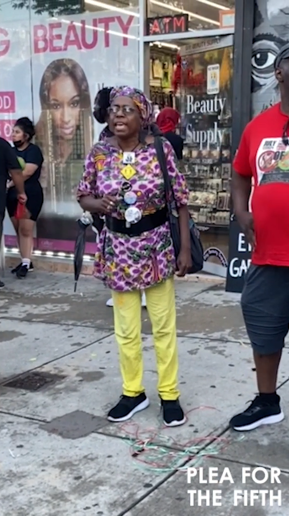 A women wearing yellow pants and a purple and black leopard print shirt in the middle of speaking. There a several buttons pinned to her t-shirt and she is wearing a headwrap.